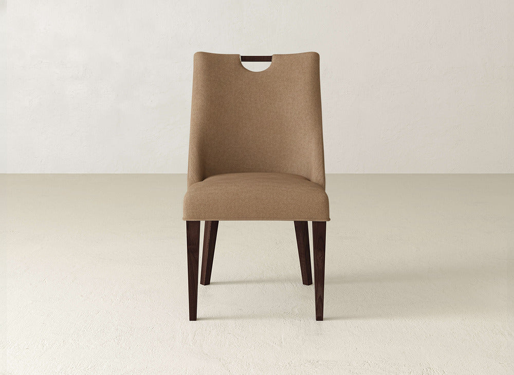 The Rosalie Dining Chair by James & James features brown upholstery, dark wood legs, and a small wooden handle cutout at the top of the backrest, shown on a light floor against an off-white wall.