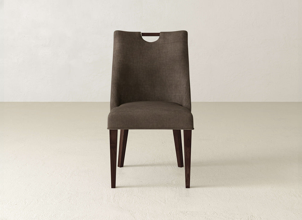 The James & James Rosalie Dining Chair features modern brown upholstery, a high back with slightly flared sides, a top cutout handle, and dark wooden legs, shown against a plain light background.