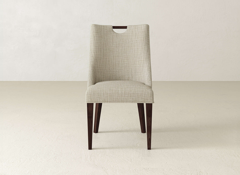The Rosalie Dining Chair by James & James offers light beige upholstery, dark wood legs, and a wooden handle cutout on the backrest, delivering a sleek modern look perfect for neutral dining spaces.