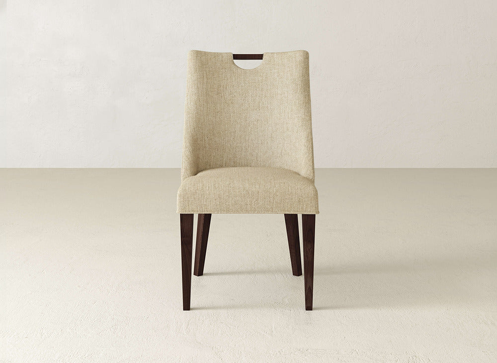 The Rosalie Dining Chair by James & James showcases beige upholstery, a modern wooden handle on the backrest, and dark wooden legs, set on a light floor with a plain, light wall background.
