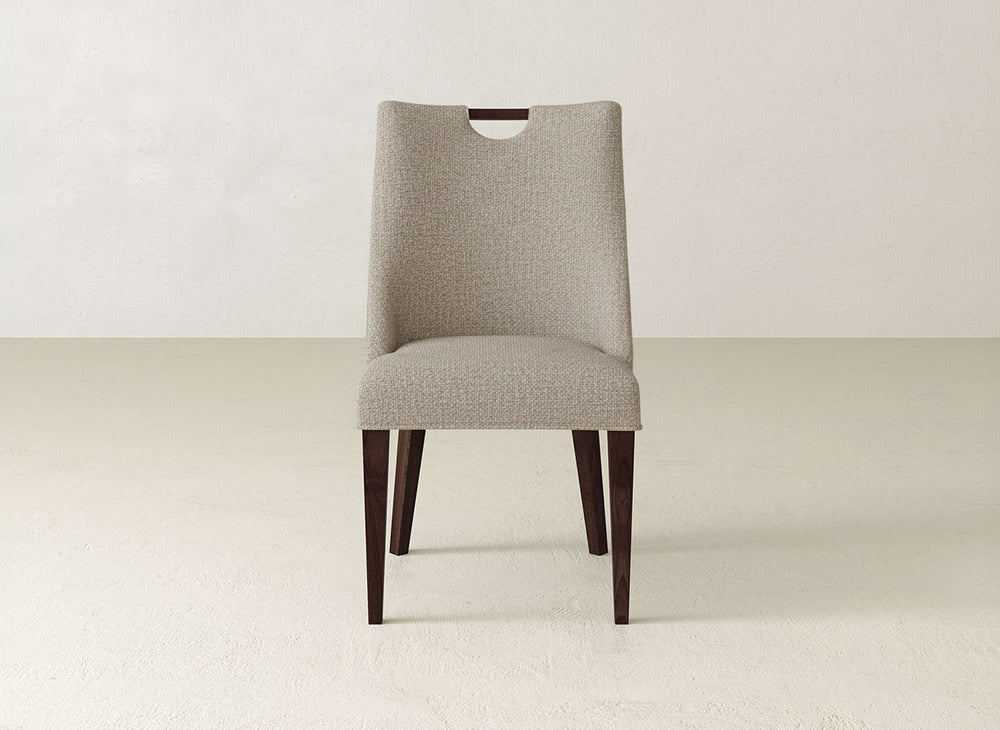 The Rosalie Dining Chair by James & James features modern beige upholstery, dark wooden legs, and a unique cut-out handle at the top, shown against a light-colored wall and floor.