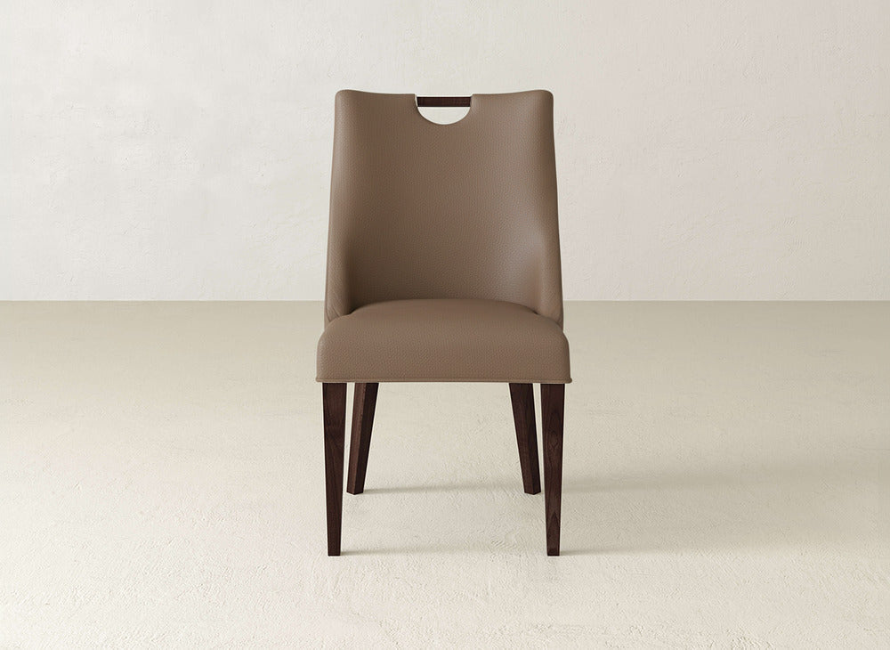 The James & James Rosalie Dining Chair features brown upholstery, dark wooden legs, and a unique handle cutout at the top of the backrest. It’s shown on a light floor with a plain light wall in the background.