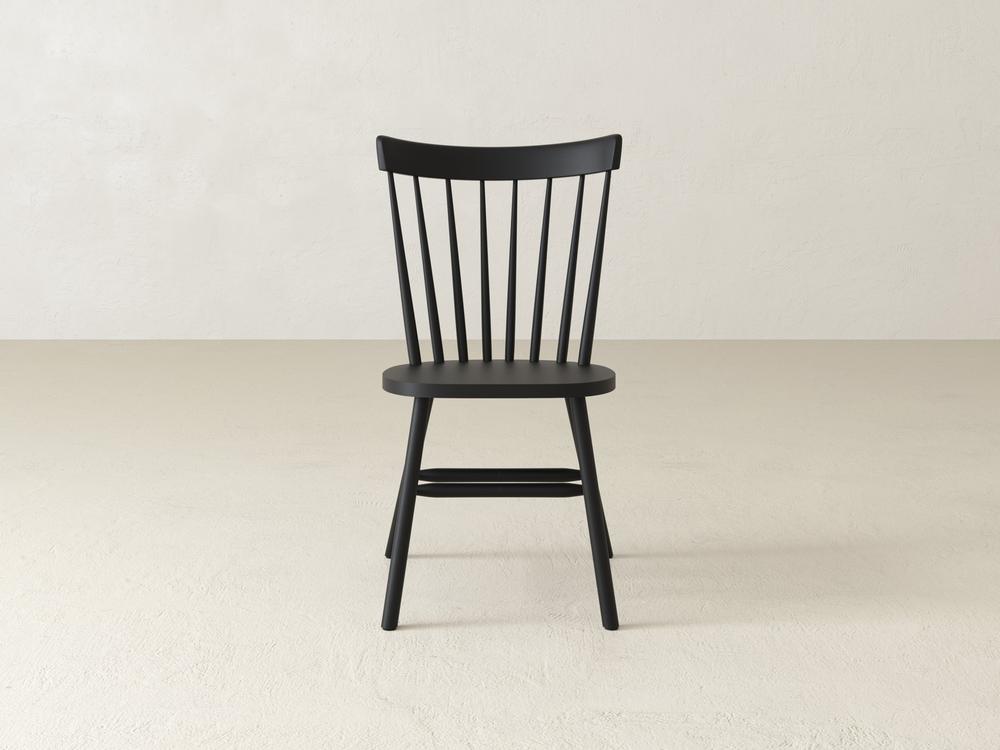 A wooden chair with a curved backrest and vertical slats sits on a light-colored floor against a plain, off-white wall. The chair has a simple and classic design.