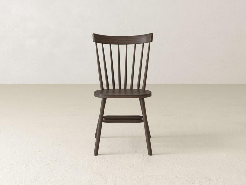 A dark wooden chair with a simple, classic design featuring a curved backrest with vertical slats, sits on a light, plain floor against a neutral wall background.