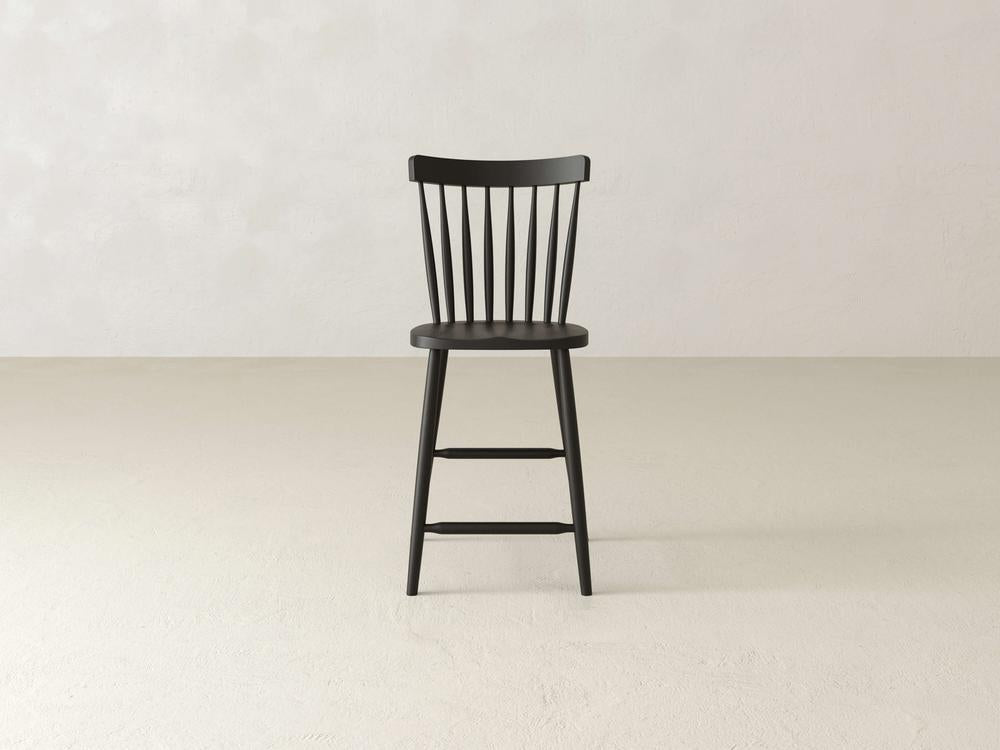 The Rustic Windsor Stool by James & James, crafted from sturdy maple hardwood, features a black finish with an elegant spindle backrest. It is shown on a light, neutral floor against a plain wall, highlighting its minimalist design and robust legs.