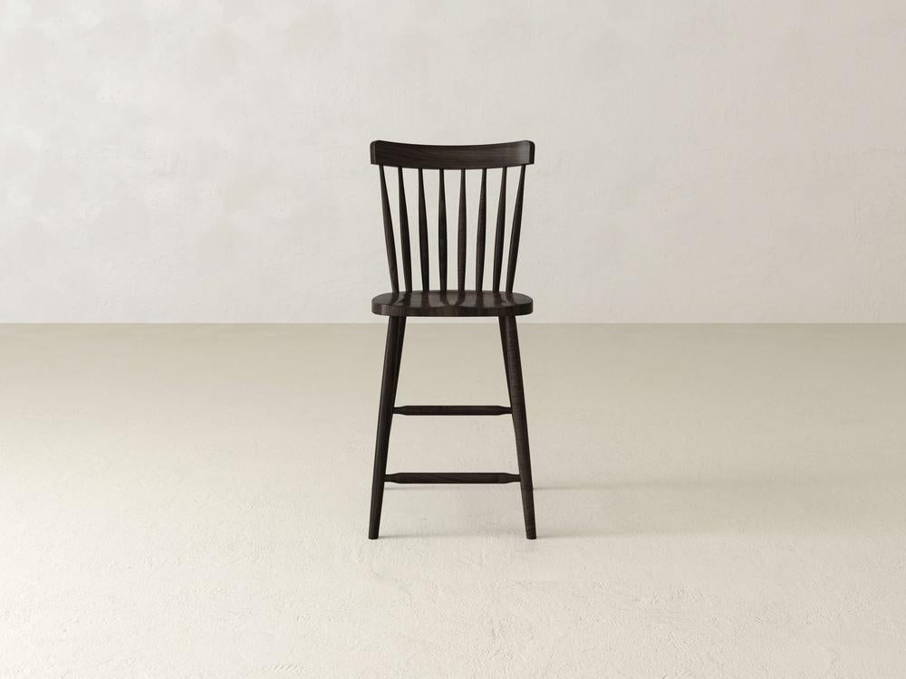 The Rustic Windsor Stool by James & James, crafted from sturdy maple hardwood, features a black finish with an elegant spindle backrest. It is shown on a light, neutral floor against a plain wall, highlighting its minimalist design and robust legs.