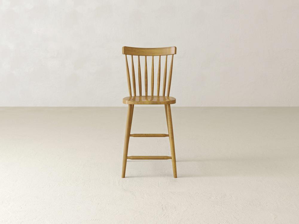 A Rustic Windsor Stool by James & James sits in a minimalist room with light walls and flooring. Made from durable maple hardwood, this wooden high-back chair features an elegant spindle backrest and a simple natural finish, positioned facing forward.