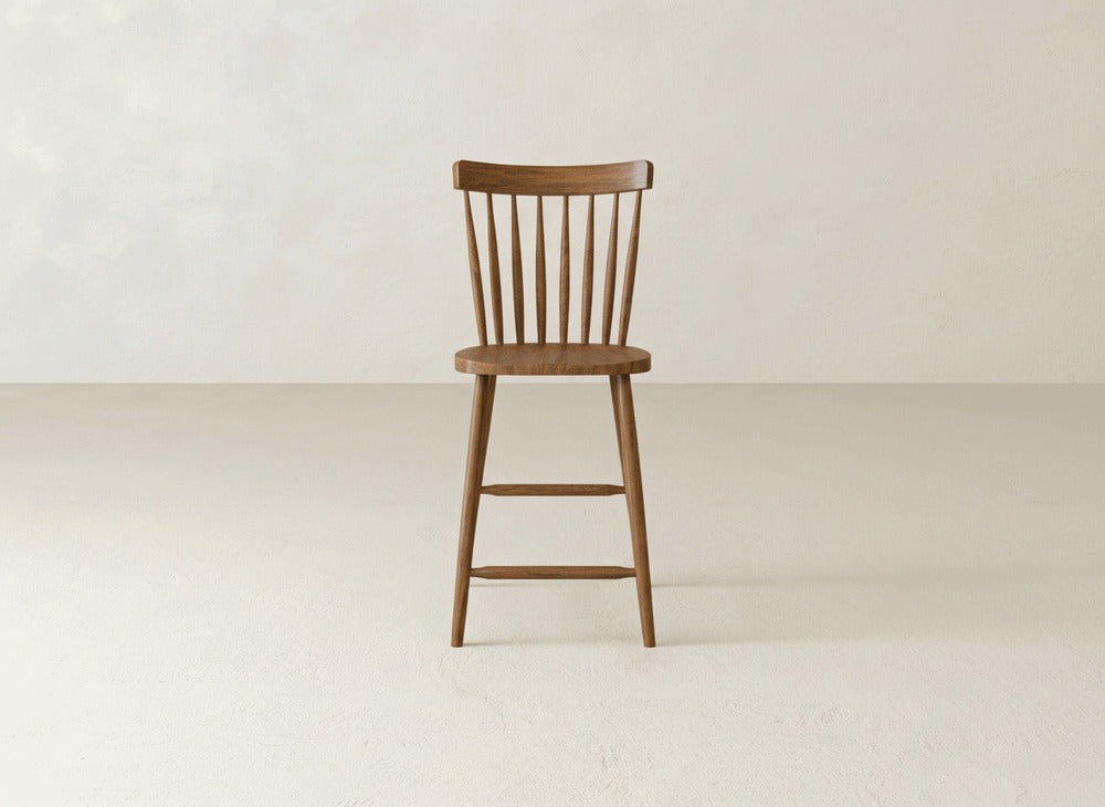 The James & James Rustic Windsor Stool, featuring a spindle backrest and vertical slats, stands on a light floor against a plain off-white wall.