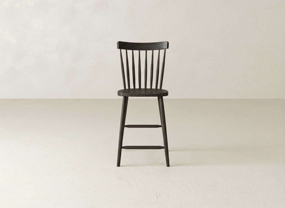 The Rustic Windsor Stool by James & James, crafted from maple hardwood with a spindle backrest, stands on a light floor against a neutral wall.