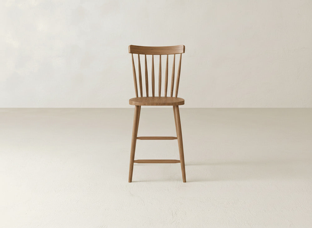 The James & James Rustic Windsor Stool, made from maple hardwood with a rounded seat, spindle backrest, and four legs linked by footrests, is placed on a light floor against an off-white wall.