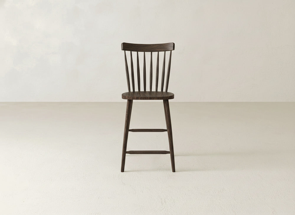 The Rustic Windsor Stool by James & James, made from maple hardwood, features a dark wood finish, curved spindle backrest, and vertical slats. It stands alone on a light floor against an off-white wall.