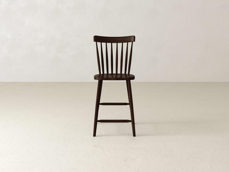 The James & James Rustic Windsor Stool, crafted from durable maple hardwood, showcases an elegant spindle backrest. It elegantly stands on a plain floor with a round seat and four legs enhanced by horizontal support bars.