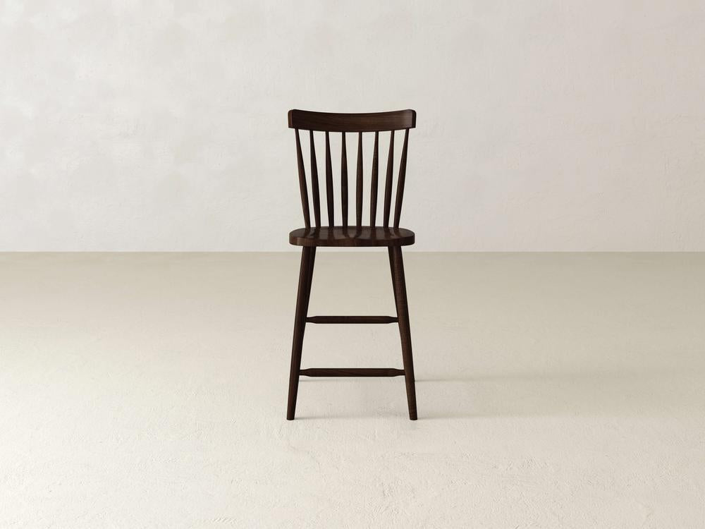 The James & James Rustic Windsor Stool, crafted from durable maple hardwood, showcases an elegant spindle backrest. It elegantly stands on a plain floor with a round seat and four legs enhanced by horizontal support bars.