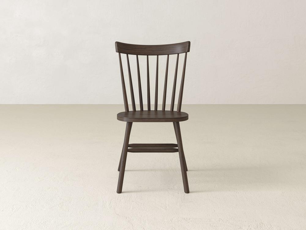 A dark wooden chair with a simple, classic design featuring a curved backrest with vertical slats, sits on a light, plain floor against a neutral wall background.