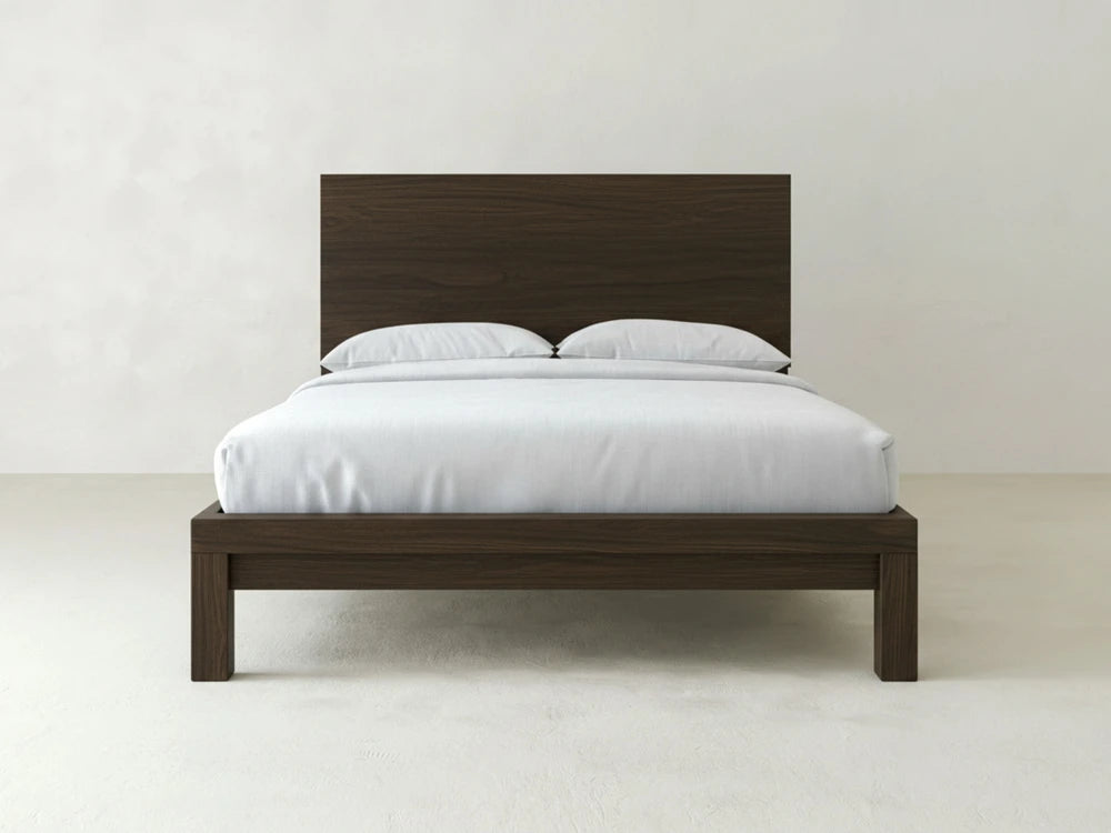 The Sawyer Bed - Espresso on Oak by James+James features a tall headboard and modern wooden frame, dressed in white bedding with two pillows, set against a plain white wall and floor.