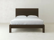 The Sawyer Bed - Espresso on Oak by James+James features a tall headboard and modern wooden frame, dressed in white bedding with two pillows, set against a plain white wall and floor.