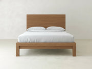 The Sawyer Bed - Almond on Oak by James+James features a minimalist wooden frame with a matching headboard, styled with white bedding and two pillows, set against a plain white wall in a bright, uncluttered room.