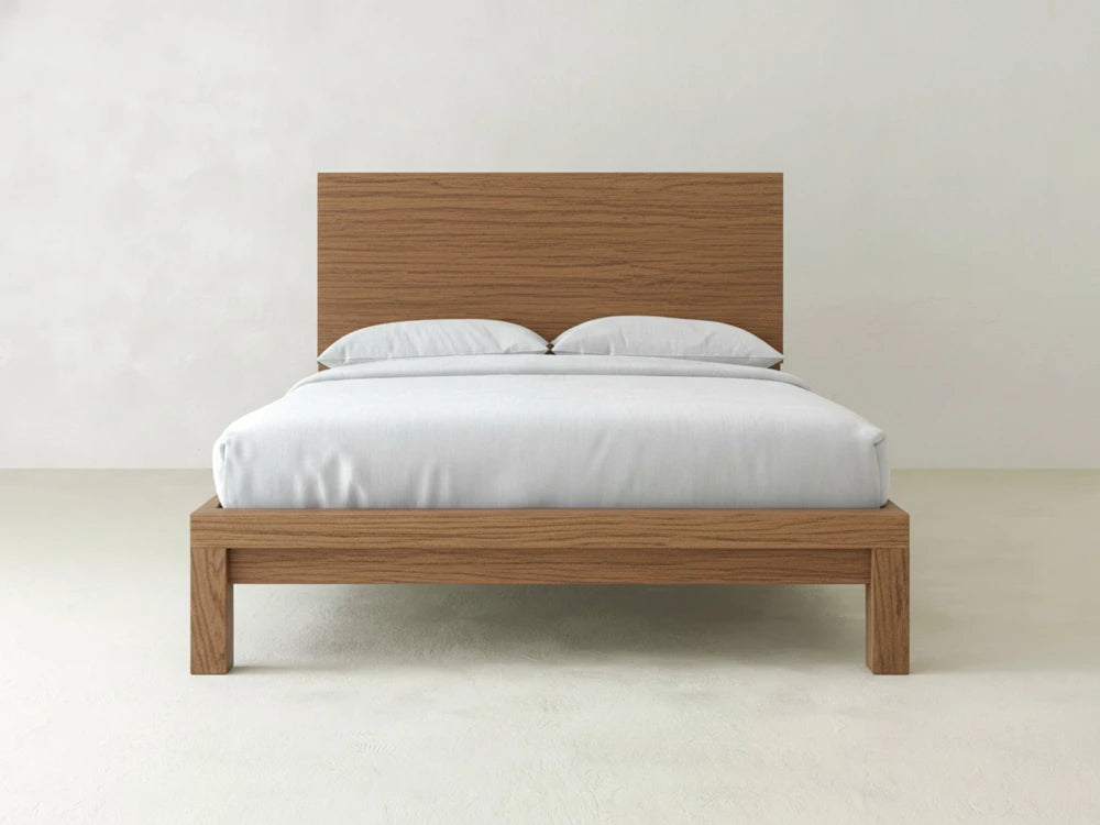 The Sawyer Bed - Almond on Oak by James+James features a minimalist wooden frame with a matching headboard, styled with white bedding and two pillows, set against a plain white wall in a bright, uncluttered room.