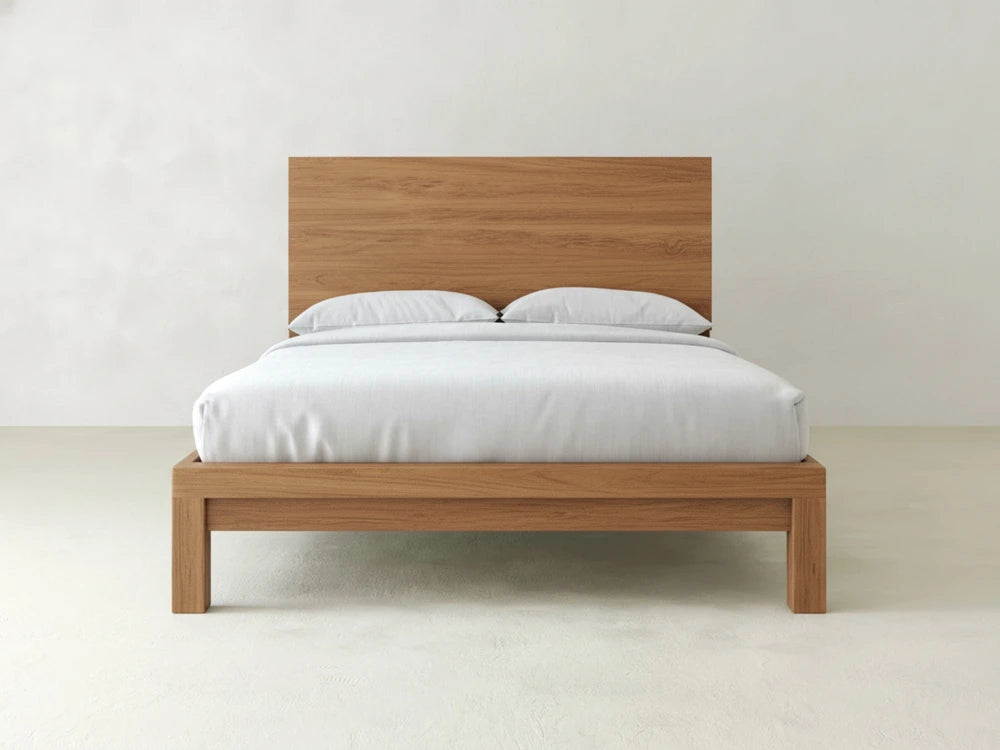 The Sawyer Bed - Oat on Oak by James+James, featuring a minimalist oak frame and simple headboard, is dressed in white bedding with two pillows and set against a plain white wall.