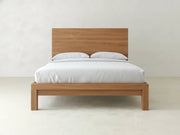 The Sawyer Bed - Oat on Oak by James+James, featuring a minimalist oak frame and simple headboard, is dressed in white bedding with two pillows and set against a plain white wall.