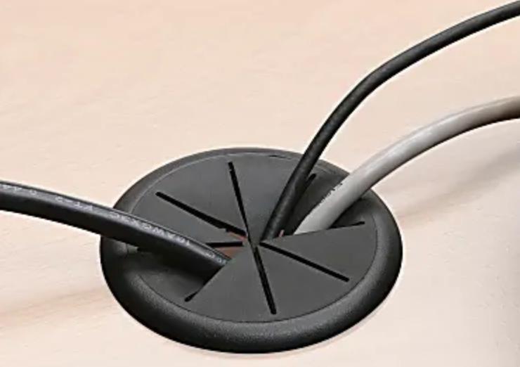 The James+James Standard Desk Add-On, a round black desk grommet with slits, accommodates three cables—black, gray, and white—effectively organizing and minimizing cable clutter on a light wood surface.