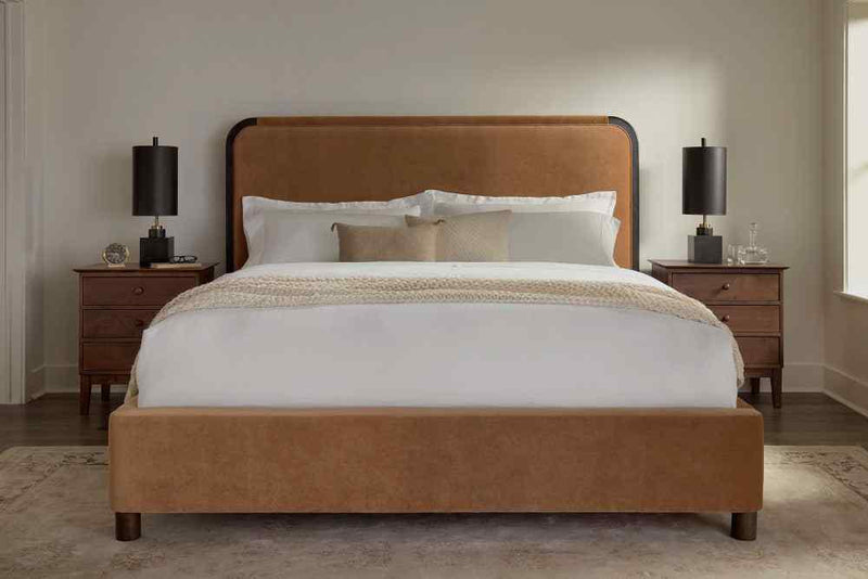 A tidy bedroom exudes modern elegance with the Sloane Bed – Linen by James & James, featuring a tan upholstered headboard, dressed in performance linen and a textured beige throw. Two matching wooden nightstands flank the bed, each with a sleek black lamp, atop a light rug on the dark wood floor.