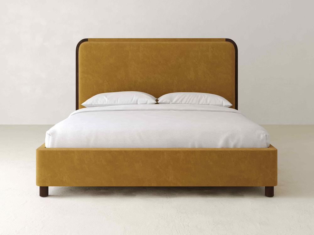 The Sloane Bed – Velvet by James & James is upholstered in mustard velvet, featuring a tall plush headboard, complemented with a white mattress and two pillows. It is positioned against a light gray wall on a matching floor.