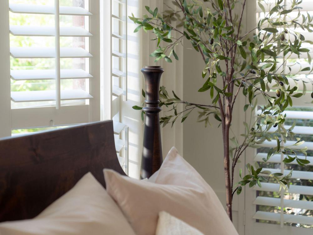 A cozy bedroom corner features the Stella Bed - Tobacco on Alder by James+James, paired with pale pillows, a leafy potted plant, and bright windows framed by white plantation shutters. Sunlight streams in, creating a serene atmosphere.