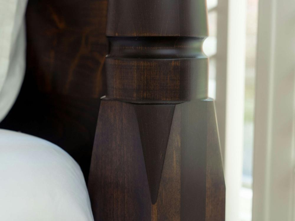 Close-up of the Stella Bed in Tobacco on Alder by James+James, featuring a dark wooden bedpost with geometric carvings beside a white bedsheet, softly illuminated by natural window light.
