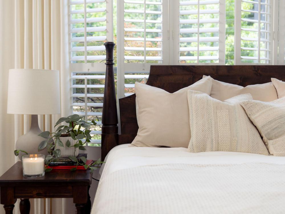 A cozy bedroom featuring the James+James Stella Bed in Tobacco on Alder, cream bedding and pillows, a bedside table with a lamp, a plant, candle, and large windows with white shutters that let in natural light.