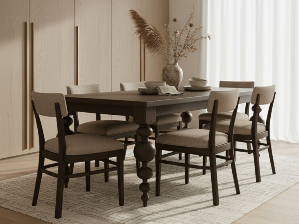 A James+James Stone dining table with six cushioned chairs in Boucle sits on a light patterned rug. Plates, cups, and a vase with dried plants decorate the table, while light streams through sheer curtains in the background.