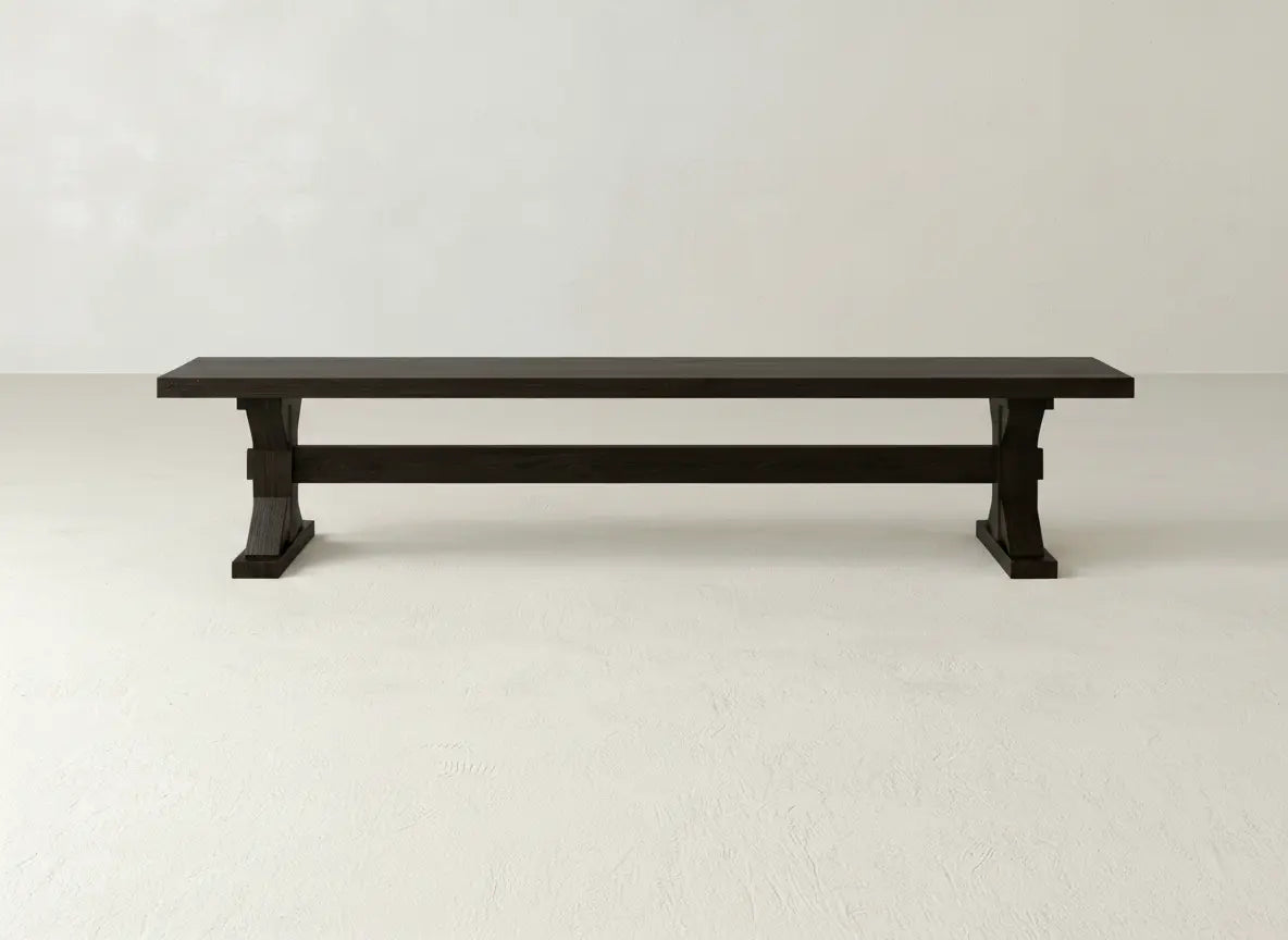 The James+James Trestle Bench in Espresso on Oak, featuring a simple and sturdy design, stands against a plain white wall on a light-colored floor.