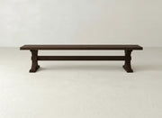 The Trestle Bench - Sable on Oak by James+James features a long, dark wooden seat with sturdy, simple legs, set against a plain, light-colored wall and floor.
