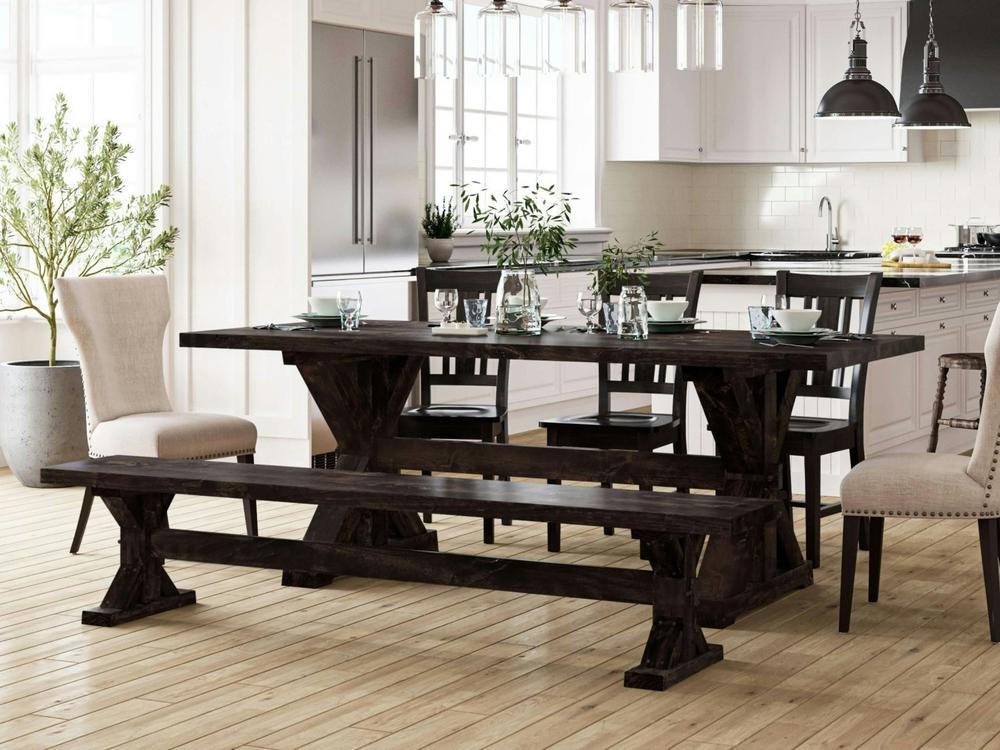 An airy, sunlit kitchen dining area features the James+James Trestle Bench in Charred Ember on Alder with a dark wood table and chairs, set with dishes and glasses. Light floors, white cabinets, plants, and pendant lights complete the modern farmhouse look.