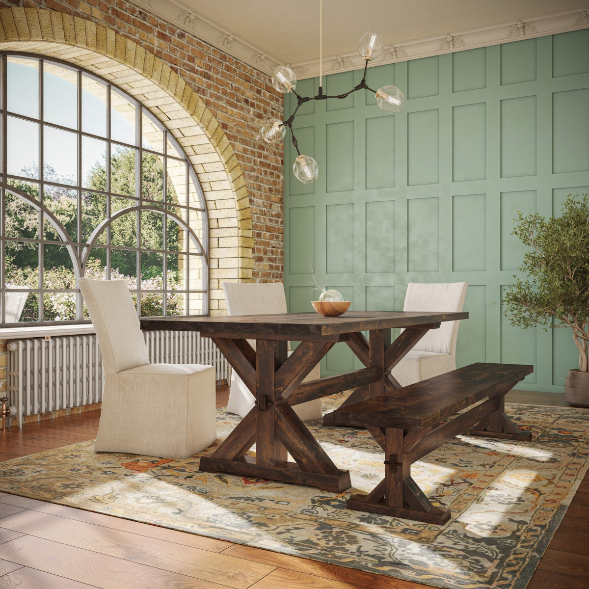 A dining room with a rustic wooden table and bench, flanked by two beige upholstered chairs. Large arched window with brick detailing. Light teal paneled wall, modern chandelier, and a small tree in the corner. Vintage-style rug on wooden floor.