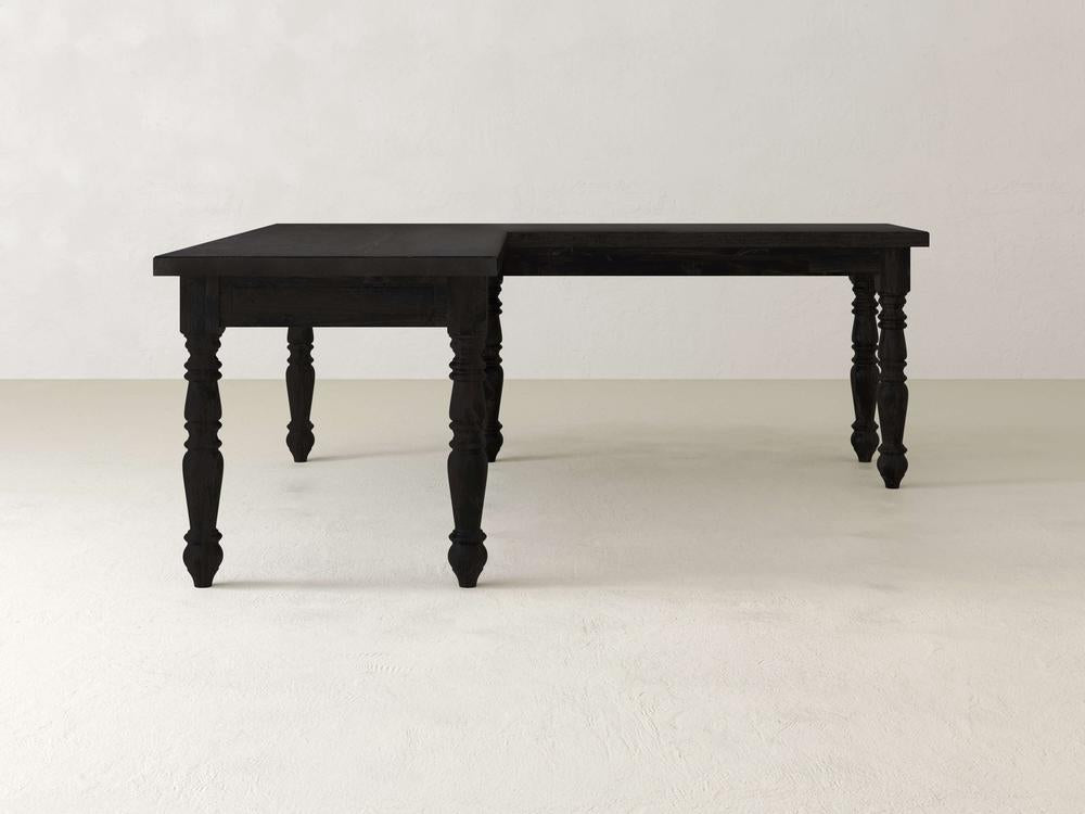 The Turned Leg Corner Desk - Charred Ember by James+James, with its black wooden L-shaped design, features ornate carved legs and a smooth surface. It stands on a plain, light-colored floor against a simple off-white background.