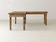 The Turned Leg Corner Desk by James+James, crafted from solid hardwood, features a distinctive L-shaped design and finely carved legs. It stands elegantly on a light-colored floor against a neutral wall, showcasing a natural wood grain finish that enhances its charm as an elegant addition to any workspace.