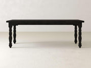 A Charred Ember Turned Leg Desk by James+James, featuring intricately carved black wooden legs, is positioned on a light beige floor against a plain white wall.