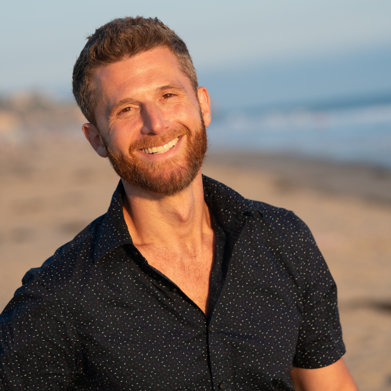 Smiling man with a beard and short hair, wearing a dark shirt, standing on a sunny beach. The ocean and sandy shore create a blurred background, suggesting a warm, serene day.