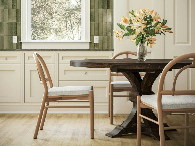 A dining area with a dark wooden round table and four light wooden chairs with white cushions. A vase with orange lilies sits on the table. There is a large window with a view of trees, and the walls are lined with green tiles above cream cabinets.
