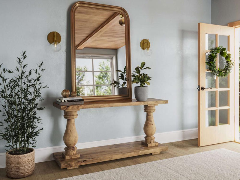 A Vivien Sofa Table in Harvest Wheat by James+James stands against a wall with a large mirror above it. Two potted plants are on the table, and a third is on the floor beside it. The doorway to the right features a wreath, and the light blue walls are adorned with two wall sconces.