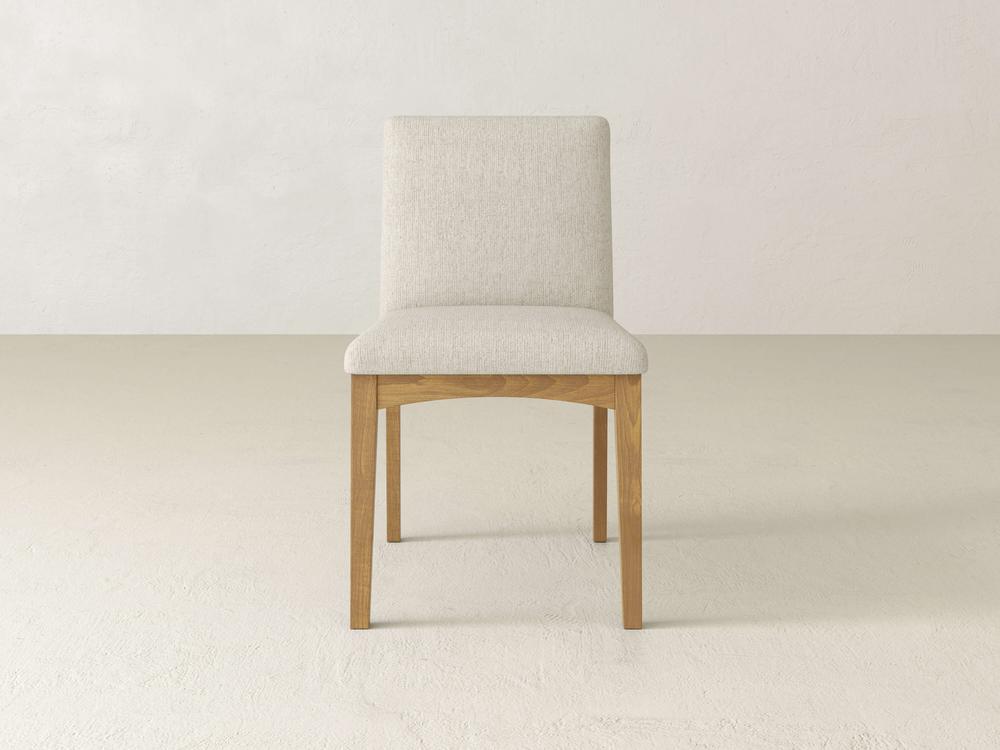 The Klein Dining Chair by James & James features a beige upholstered back and light wooden frame. Its minimalist design with elegant maple legs rests on a neutral floor, set against a plain white wall.