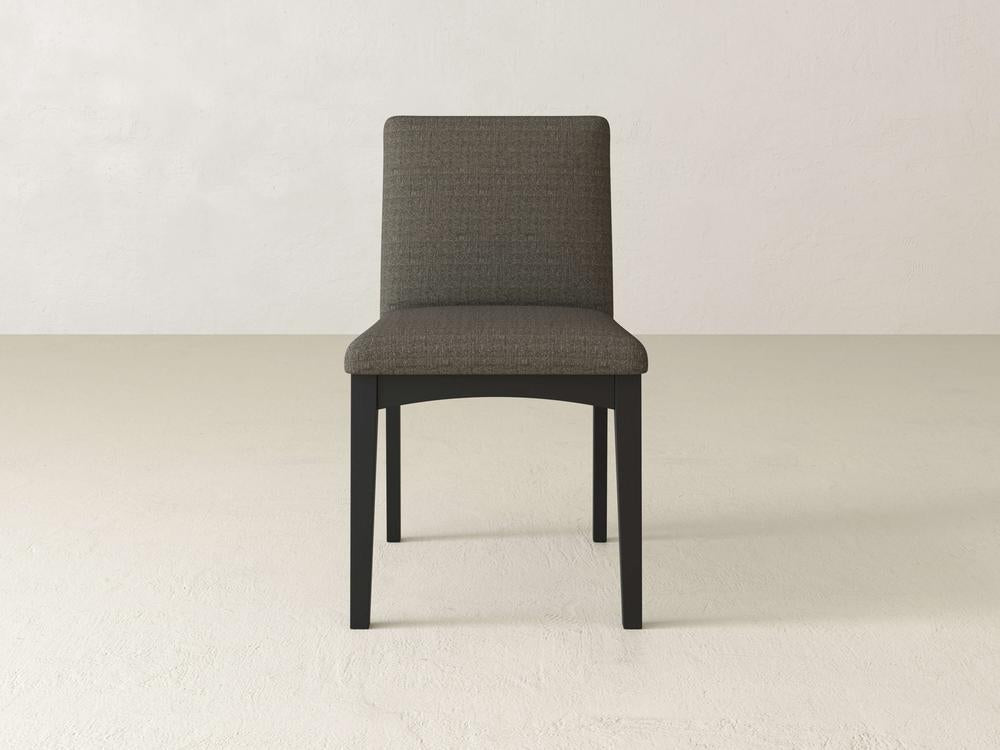 The James & James Klein Dining Chair showcases a modern design with an upholstered, padded back in dark gray fabric. It features a black wooden frame and maple legs, beautifully set on a light-textured floor against a plain white wall.