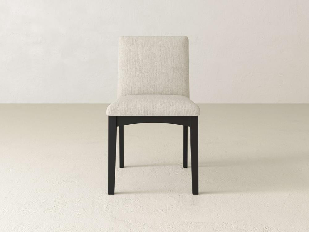 The Klein Dining Chair by James & James, with its upholstered back and maple wooden legs in beige, stands gracefully on a light-colored floor, contrasting elegantly against the plain white wall.
