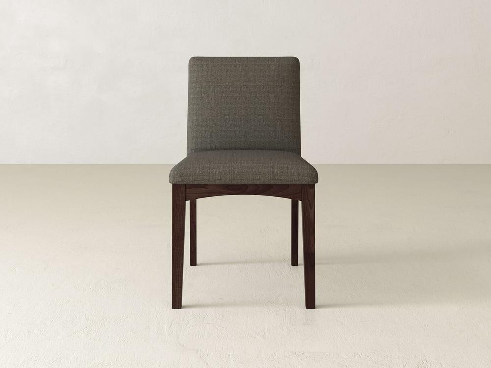 The Klein Dining Chair by James & James, featuring maple wooden legs and an upholstered back, stands against a plain, light-colored wall and floor. Its gray cushioned seat and backrest add comfort and style to any setting.