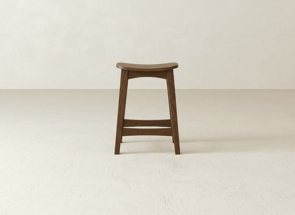 The Weston Stool by James & James features a contoured maple hardwood seat and clean lines, highlighting natural wood grain as it stands on a light textured floor against an off-white wall.