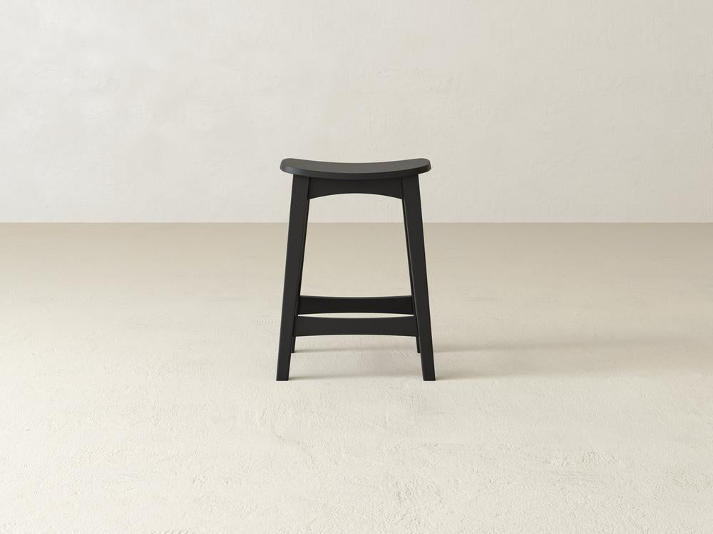 A simple Weston Stool by James & James features a contoured seat and straight legs in black maple hardwood, set on a light beige floor against a white textured wall.