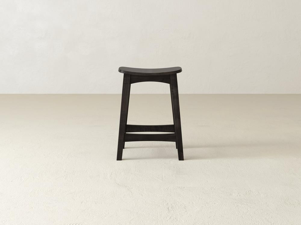A simple Weston Stool by James & James features a contoured seat and straight legs in black maple hardwood, set on a light beige floor against a white textured wall.