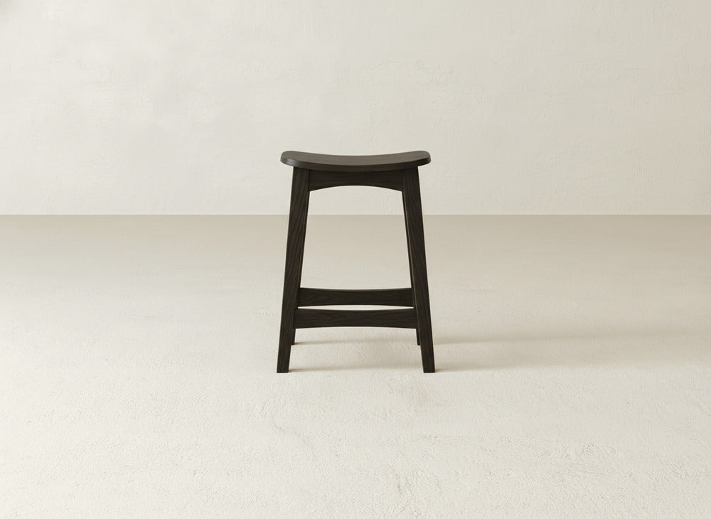 The Weston Stool by James & James features a contoured dark maple hardwood seat, clean lines, visible natural wood grain, and no backrest. It stands on a plain light floor against an off-white wall.