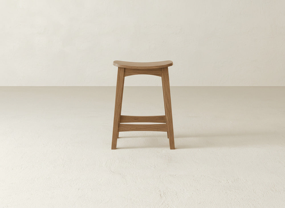 The Weston Stool by James & James features a contoured maple hardwood seat and footrest, highlighting natural wood grain. It stands on a light-textured floor with an off-white wall as a backdrop.